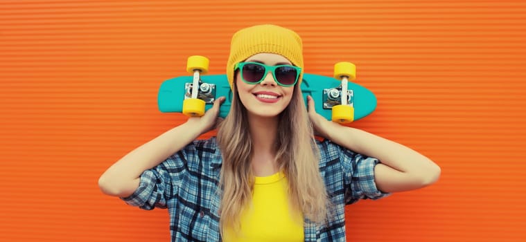 Summer portrait of happy cheerful stylish young woman with skateboard in colorful clothes, yellow hat posing on orange background on city street