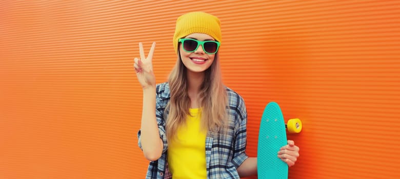 Summer portrait of happy cheerful stylish young woman with skateboard in colorful clothes, yellow hat posing on orange background on city street