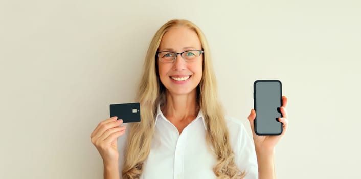 Portrait of happy smiling middle-aged woman holding plastic credit bank card and blank screen phone on white studio background