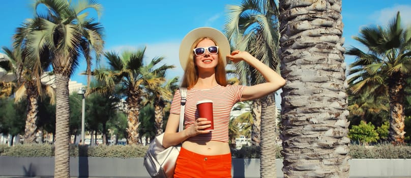 Beautiful young woman with cup of coffee in summer park wearing backpack, straw hat, shorts against a palm trees background