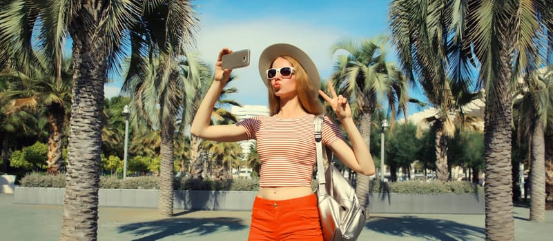 Portrait of young woman taking selfie with phone in summer park wearing backpack, straw hat against a palm trees background