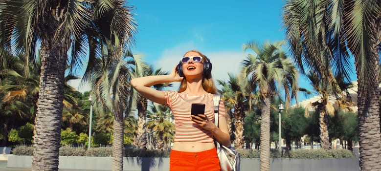 Happy smiling young woman listening to music in headphones with smartphone in summer park against a palm trees background