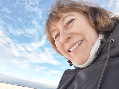 A cheerful middle aged woman in a winter coat taking selfie on nature outdoors in sunny day with blue sky