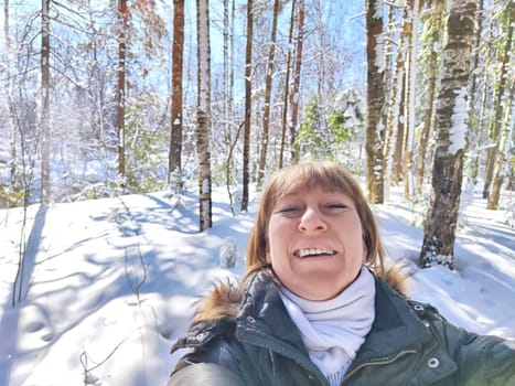 A cheerful middle aged woman in a winter coat taking selfie on nature outdoors in sunny day with blue sky