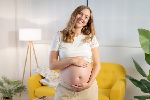 portrait of a smiling pregnant woman embracing belly in living room with copy space for text. High quality photo