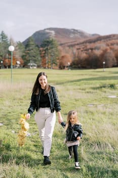 Smiling mother with a wreath of yellow leaves in her hand walks with little girl across the lawn holding hands. High quality photo