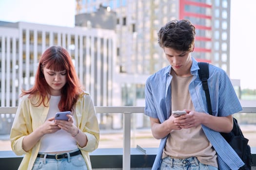 Teenage friends university college students guy and girl using smartphones, urban outdoor, city modern buildings background. Technology, friendship, youth 19-20 years old, lifestyle concept