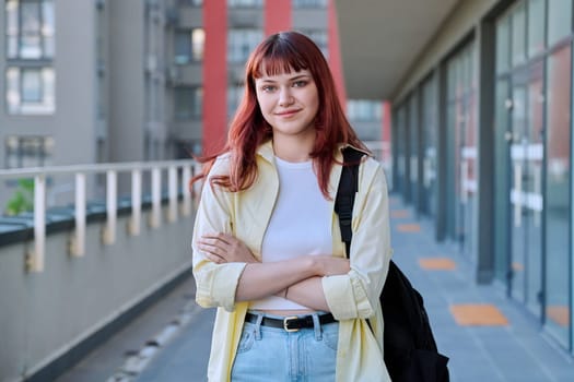 Urban portrait of young confident red-haired female with backpack with crossed arms, city outdoor. Lifestyle, youth 19-20 years old concept