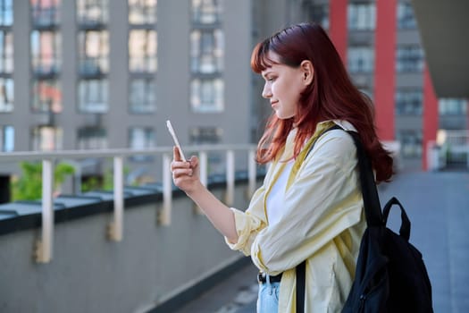 Young female using smartphone, modern city background. Teenage student 19-20 years old, texting using mobile apps applications for leisure study travel. Technology Internet education youth urban style