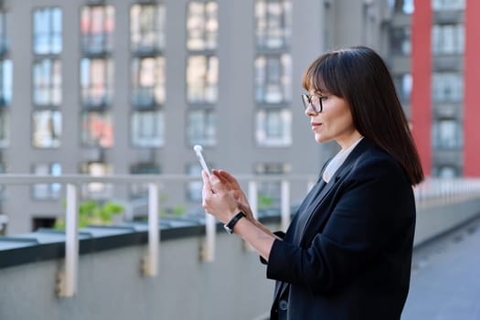 Mature business confident female using smartphone, modern city background. Middle aged successful woman texting using mobile applications apps for business work. Technology internet job, urban style