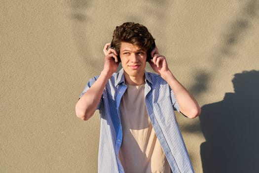 Young handsome male in wireless headphones listening to music, against the backdrop of an outdoor sunny gray wall