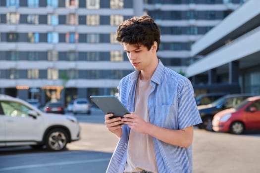 Young male using digital tablet on city street. Handsome guy student 19-20 years old, using pad for leisure, studying, working. Technology, youth, education, urban style concept