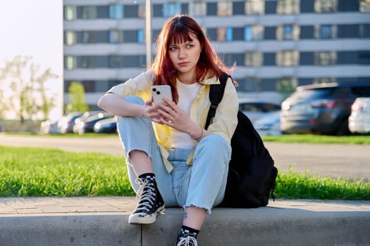 Young beautiful red-haired woman using smartphone, sitting on floor, sunny day outdoor city urban style. Youth, lifestyle, technology, internet online applications for leisure tourism learning work