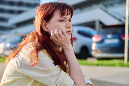 Upset sad unhappy young female sitting on steps. Pensive serious beautiful red-haired girl student 19-20 years old sitting outdoor. Problems, difficulties, depression, mental health of young people