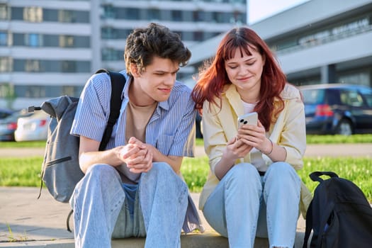 Friends young guy and girl university college students talking together, looking at smartphone outdoor, city modern buildings background. Lifestyle, communication, youth 19-20 years old, urban style