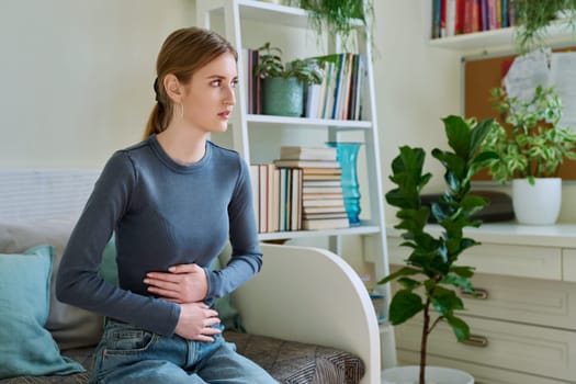 Young teenage female experiencing abdominal pain sitting at home. Stomach, menstrual cramps, gastrointestinal tract diseases, inflammatory processes in abdominal organs, kidneys, pelvis, youth, health