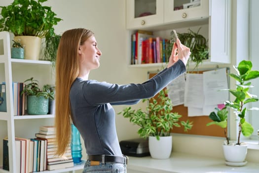 Fun grimacing happy young teenage female taking selfie photo on smartphone, in home interior. Joy, fun, youth, lifestyle concept