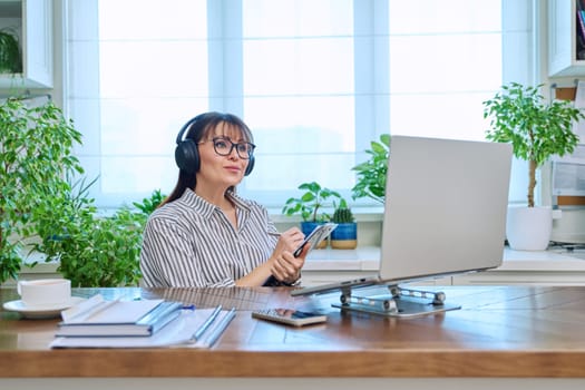 Mature woman in headphones having video conference using laptop computer in home office. Remote virtual meeting, online training, consulting, testing. Work, education, technology, people concept