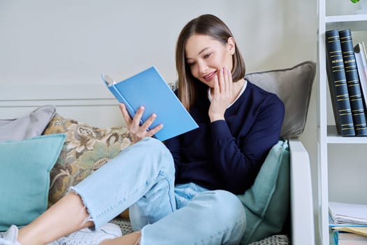 Young woman reading paper book, relaxed sitting on sofa at home. Fiction, hobby, leisure, youth concept