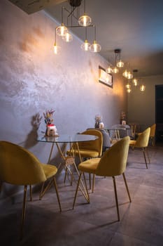 Cozy restaurant interior with glass tables, yellow chairs, textured walls, and pendant lights hanging from the ceiling.