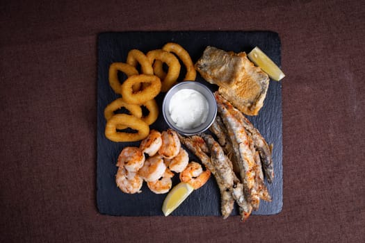 A delicious and healthy meal of fried seafood, including shrimp, fish, and calamari, served with lemon wedges on a black slate plate.