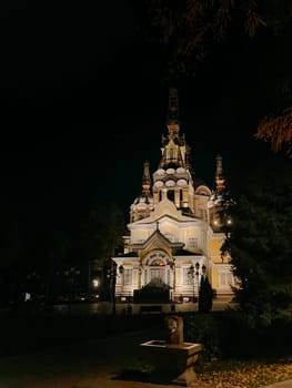 A stunning Russian Orthodox church illuminated at night, with a vibrant yellow exterior, towering steeple, surrounded by trees and park.