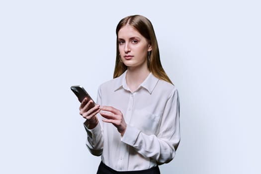 Teenage student girl 16, 17, 18 years old in white shirt holding smartphone in hands, serious looking at camera on white studio background. Education, technology, high school, college, lifestyle, youth concept