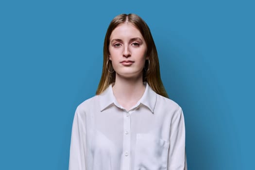 Portrait of young teenage smiling female in white shirt on blue studio background. Confident beautiful happy college student girl looking at camera