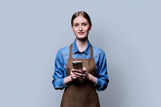 Young female worker in an apron looking at camera holding smartphone on gray studio background. Work, business, online internet services, mobile apps applications, technology concept