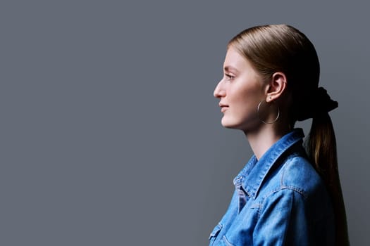 Profile view, young serious confident female looking forward on gray studio background, copy space for advertising text image. Beautiful teenage blonde with long hair in denim shirt, fashion style beauty youth