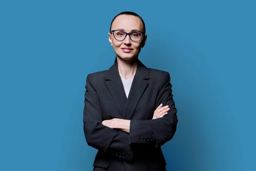 Portrait of business woman in 30s with crossed arms, on blue studio background. Confident female in glasses, suit looking at camera. Business, work, job, career, people concept