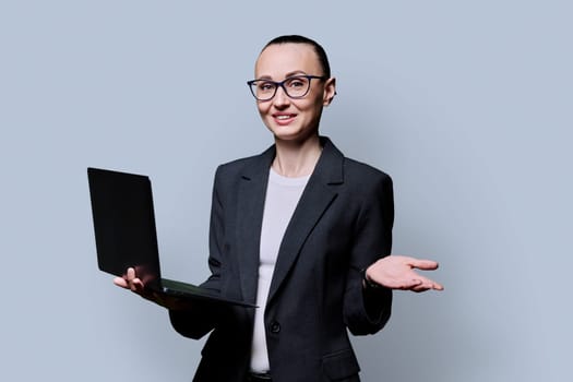 Middle-aged business serious woman using laptop on gray background. 30s successful female teacher mentor manager worker employee director looking at camera. Internet online technologies work teaching