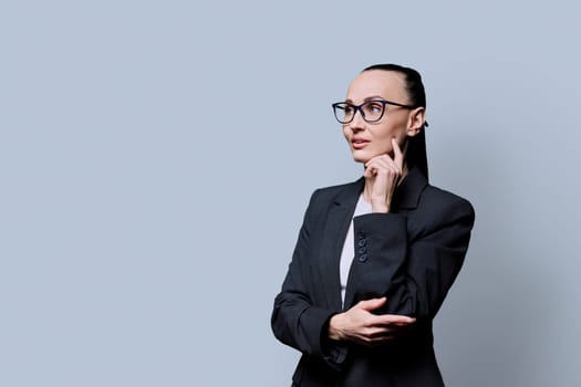 Portrait of thinking serious 30s business woman on grey studio background. Confident female in glasses suit looking to side at space for image text copy space. Business work teaching job career people
