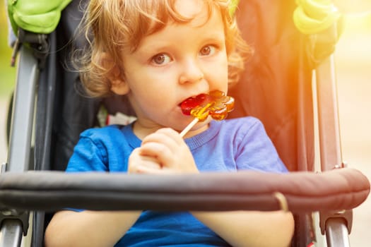 Toddler in blue shirt eating lollipop in stroller, sunny day outdoors. Casual snapshot for design and poster.
