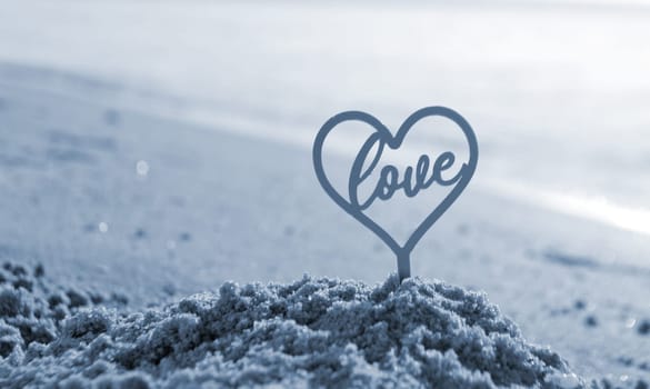 Plastic stick in shape of heart and word Love in sand on beach seashore on sunny summer day close-up. Figures in shape of heart word Love on sea waves. Love relationship romance concept. Blue color