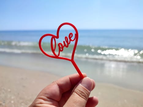 Person holding in fingers hand stick in shape red heart and word Love on background sea and sea waves, blue sky in seashore on sunny summer day close-up. Concept love romance amour St Valentines Day