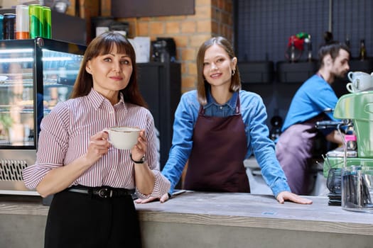 Woman customer of coffee shop near counter with cup of coffee looking at camera, restaurant workers owners at workplace. Colleagues partners in food service work, entrepreneurship, small business