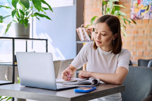 Young female college student studying using laptop, writing in notebook while sitting in coworking cafe. E-learning, education, lifestyle, youth concept