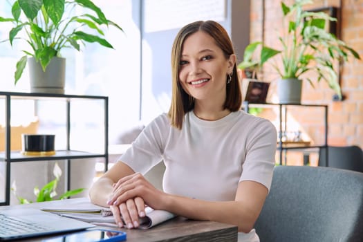 Portrait of beautiful smiling young woman sitting at table in coworking cafe, stylish pretty female 20s student looking at camera. Youth, beauty, university college students, lifestyle concept