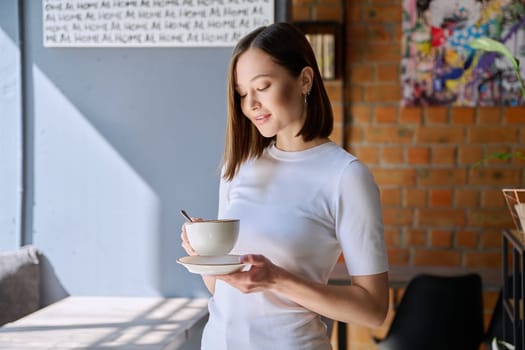 Young beautiful happy woman holding cup of coffee tea with saucer looking at cup, gray wall of cafe coffee shop cafeteria copy space for text. Coffee business work services youth lifestyle leisure