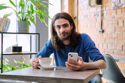 Young 30s handsome bearded with long hair man using smartphone drinking cup of coffee. Smiling guy looking at camera sitting in cafe. Mobile technologies apps for leisure work business communication