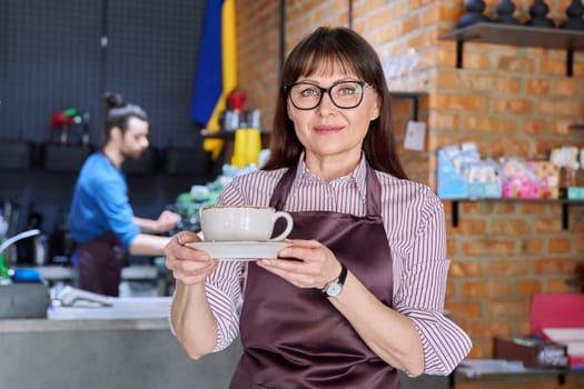 Woman in apron, food service coffee shop worker, small business owner with cup of fresh coffee, looking at camera near bar counter with male barista. Staff, occupation, entrepreneur, work concept