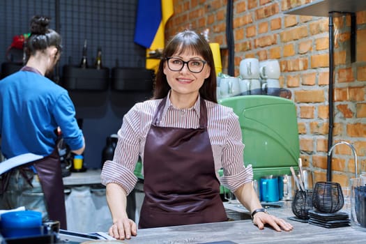 Portrait of 40s female small business owner entrepreneur in apron standing behind bar counter in coffee shop cafeteria cafe. Staff partnership teamwork work service retail management success