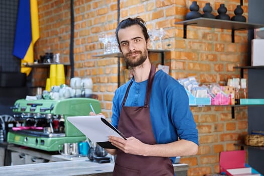 Young man in apron, food service worker, small business owner entrepreneur with work papers looking at camera near counter of coffee shop cafe cafeteria. Staff, occupation, successful business, work