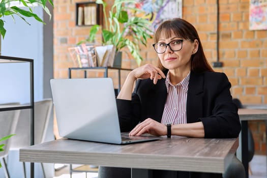 Middle-aged business confident woman working remotely at table with laptop computer looking at camera in coworking cafe. Business, mature people, success, leadership, management, empowerment concept