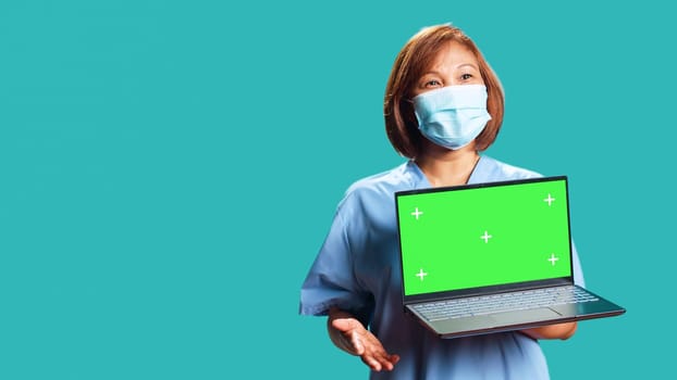 Close up handheld camera shot of happy clinic worker showing instructions video on laptop chroma key green screen. Nurse isolated over studio background presenting healthcare tape on gadget