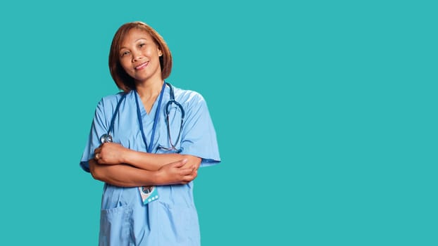 Joyful experienced asian nurse folding arms, smiling while looking at camera. Portrait of happy healthcare expert wearing protective scrubs, isolated over blue studio background