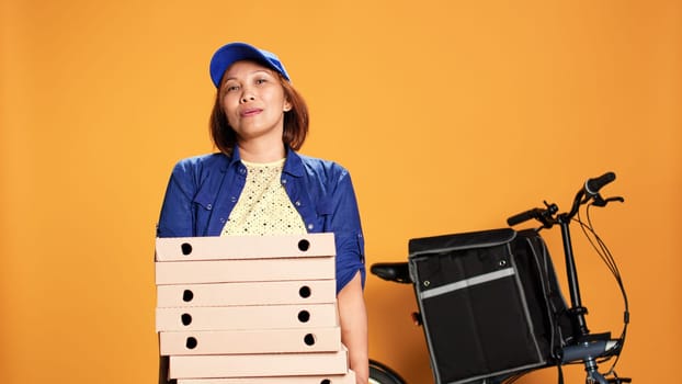 Impatient asian worker waiting for customer to answer door and pick up food. Portrait of bored courier isolated over orange studio background carrying pile of pizza boxes