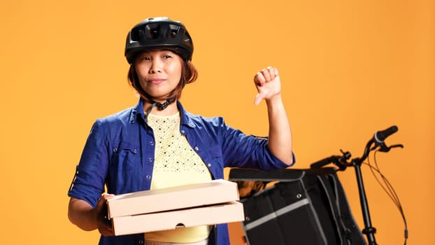 Close up shot of part time courier holding takeaway pizza boxes, showing thumbs down sign. Food delivery cyclist doing disapproving finger shaking gesture, saying no, isolated over studio background
