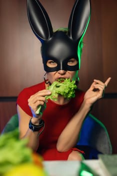 A beautiful girl in a bdsm rabbit mask and a bright red dress eats lettuce leaves promoting a healthy lifestyle and vegetarianism